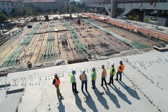 Seven construction workers in safety vests stand on a concrete deck at a building site