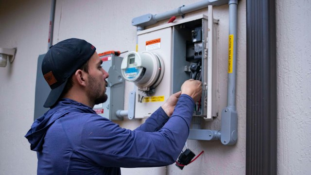 A technician in a navy hoodie and cap works on an electric meter