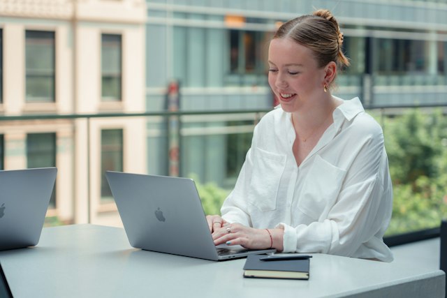 A woman in a white shirt works on a laptop at a table by a window